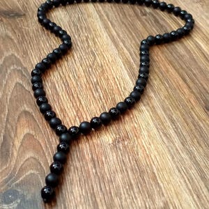 Puede incluir: Un collar de cuentas negro con un colgante en forma de Y. El collar presenta cuentas negras mate y brillantes, creando un contraste en la textura. Las cuentas están ensartadas, formando un accesorio largo y elegante.