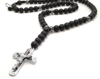 May include: A black beaded necklace with a silver cross pendant. The necklace features matte black beads interspersed with metallic gray beads. The silver cross depicts a figure and is detailed with intricate designs. The necklace is displayed on a white background.