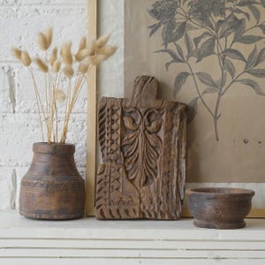 Puede incluir: Un jarrón de madera marrón oscuro con hierba seca beige, una tabla de madera rectangular con intrincados tallados de hojas y patrones geométricos, y un pequeño cuenco de madera marrón oscuro, dispuestos como un conjunto de objetos decorativos.