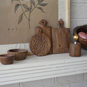 May include: A collection of rustic wooden kitchenware, including carved cutting boards, small bowls, and a utensil holder with a gold spoon. A botanical print in a wooden frame is in the background. The items are arranged on a white shelf.