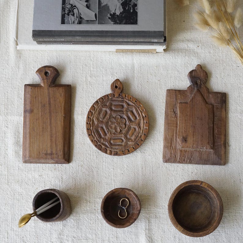 May include: An overhead shot of various wooden kitchen items. Includes three small cutting boards, two small bowls, and a small cup with a spoon and chopsticks. The items are arranged on a white surface.