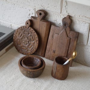 May include: A collection of rustic wooden kitchenware, including carved cutting boards, small bowls, and a cup with a gold spoon. The cutting boards have unique shapes and designs. The items are made of dark brown wood.