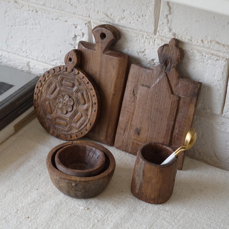 May include: A collection of rustic wooden kitchenware, including carved cutting boards, small bowls, and a cup with a gold spoon. The cutting boards have unique shapes and designs. The items are made of dark brown wood.