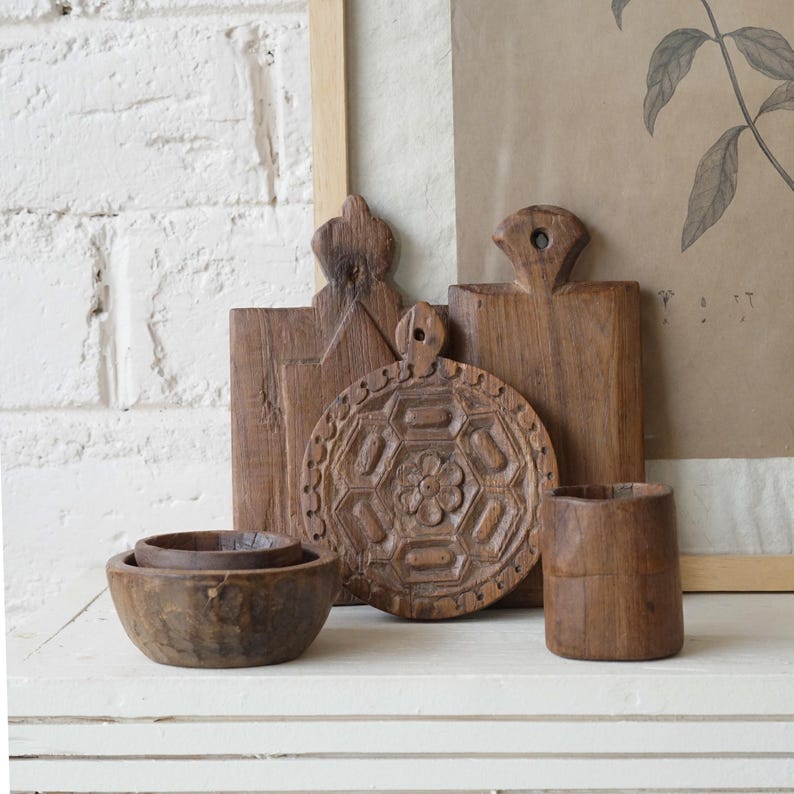 May include: A collection of handcrafted wooden kitchenware, including cutting boards, bowls, and a small cup. The items are made of a warm brown wood and have unique carved details. The arrangement is on a white surface against a white brick wall.