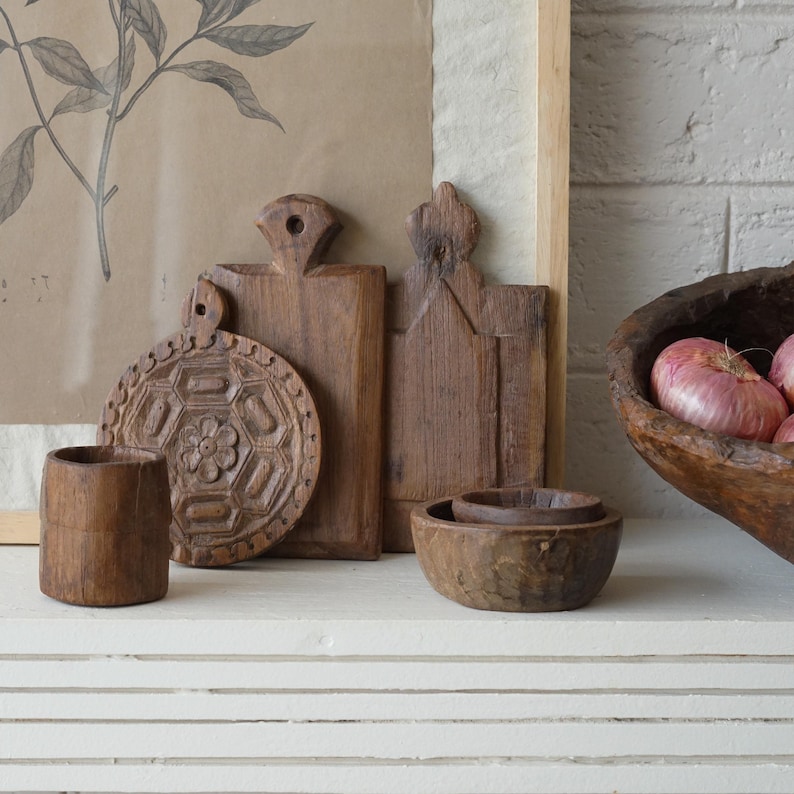 May include: A collection of rustic wooden kitchenware, including cutting boards, bowls, and a small cup. The cutting boards have unique shapes and carved details. A wooden bowl holds several red onions. The items are arranged on a white surface.