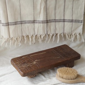 Puede incluir: Una plataforma rectangular de madera con detalles tallados y bordes redondeados. La plataforma descansa sobre una superficie blanca con una toalla a rayas marrones y blancas detrás. Un cepillo de madera con cerdas naturales está en primer plano.