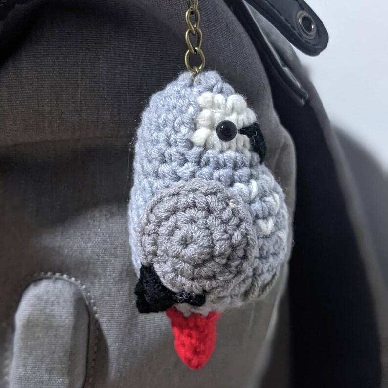 May include: A gray crocheted bird keychain with black eyes, white accents, and a red crocheted foot. The keychain is attached to a gray and black backpack.