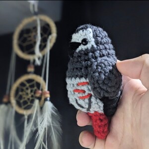 May include: A crocheted gray and black parrot with red accents. The parrot is holding a red tail feather.