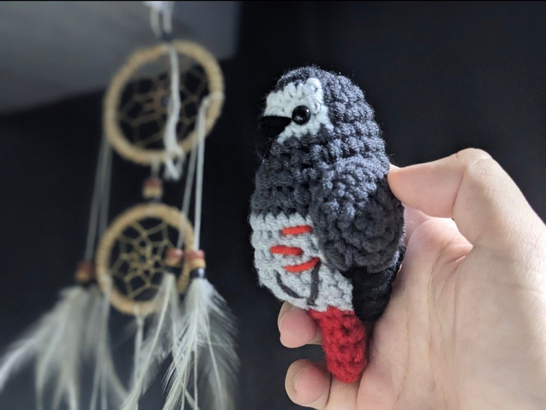 May include: A crocheted gray and black parrot with red accents. The parrot is holding a red tail feather.