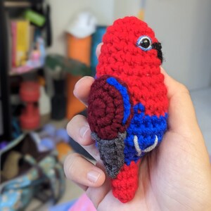 Op de afbeelding: Een gehaakt speelgoedpapegaai van rood, donkerrood, blauw en grijs garen. De papegaai heeft een zwarte snavel en &eacute;&eacute;n zwart oog.
