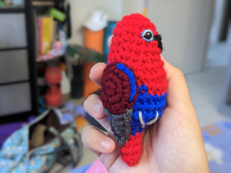 Op de afbeelding: Een gehaakt speelgoedpapegaai van rood, donkerrood, blauw en grijs garen. De papegaai heeft een zwarte snavel en &eacute;&eacute;n zwart oog.