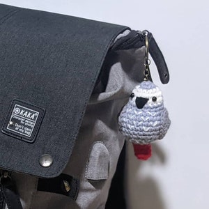 May include: A gray and white crocheted bird keychain with red feet attached to a black and gray backpack. The keychain has black eyes and a black beak. The backpack has a black tag with the text "KAKA Character design for young Don't stare at my bag".