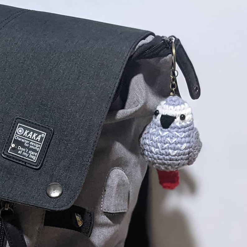 May include: A gray and white crocheted bird keychain with red feet attached to a black and gray backpack. The keychain has black eyes and a black beak. The backpack has a black tag with the text "KAKA Character design for young Don't stare at my bag".