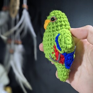 Op de afbeelding: Een gehaakt speelgoedpapegaai met groene, rode en blauwe accenten. De papegaai houdt een klein takje in zijn bek.