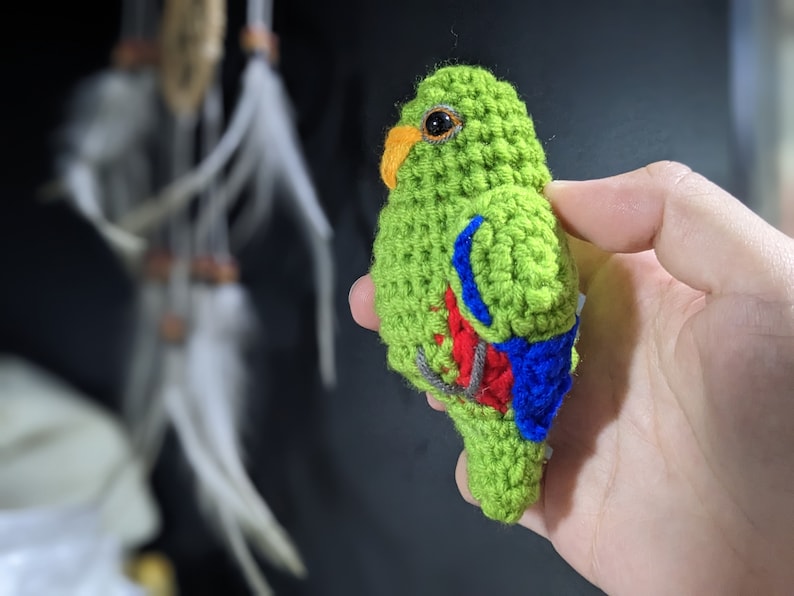 Op de afbeelding: Een gehaakt speelgoedpapegaai met groene, rode en blauwe accenten. De papegaai houdt een klein takje in zijn bek.