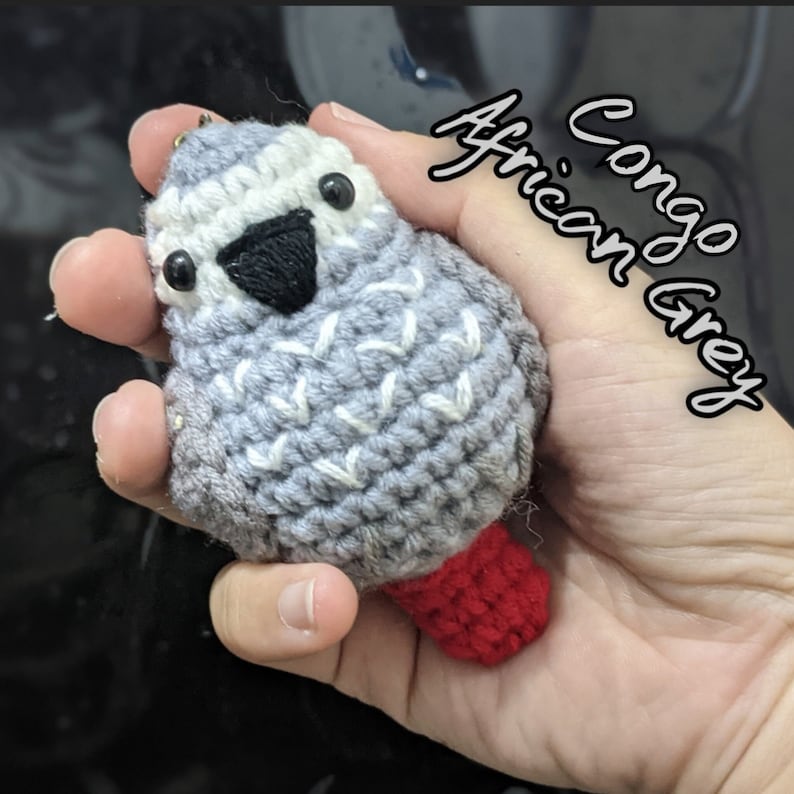 May include: A crocheted gray and white Congo African Grey parrot with a red crocheted foot. The parrot is holding a small metal ring in its beak.