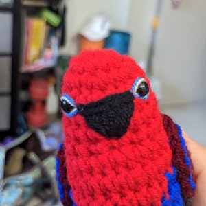 Op de afbeelding: Een rood gebreid knuffeldier in de vorm van een vogel met zwarte ogen, een zwarte snavel en blauwe en donkerrode accenten.