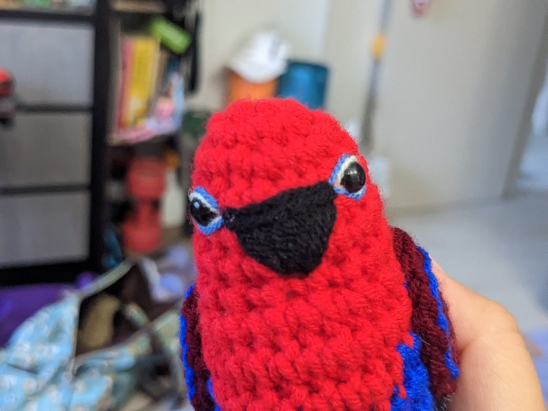 Op de afbeelding: Een rood gebreid knuffeldier in de vorm van een vogel met zwarte ogen, een zwarte snavel en blauwe en donkerrode accenten.