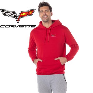 May include: A red Corvette hoodie with a front pocket and a hood. The Corvette logo is displayed on the upper left, and a smaller logo is on the chest. The person is wearing grey sweatpants.