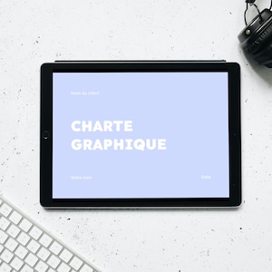 Peut inclure: Une tablette blanche avec une bordure noire affichant un écran bleu clair avec le texte "CHARTE GRAPHIQUE" en blanc. La tablette est sur une surface blanche avec un clavier blanc et des écouteurs noirs.