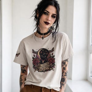 Può includere: Una t-shirt beige a maniche corte con una stampa grafica di un gatto nero circondato da rose rosse, farfalle e una luna crescente. Il testo recita: NOT YOUR BABE, NOT YOUR PROBLEM, JUST MY CAT & I. Una piccola farfalla è stampata sulla manica.