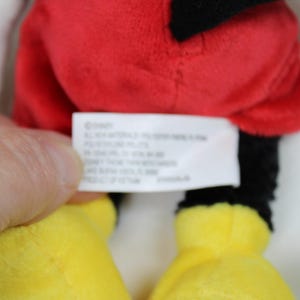 May include: Close-up of a Mickey Mouse plush toy. The toy features a red shorts, black legs, yellow feet, and white gloves. A tag with text is visible.