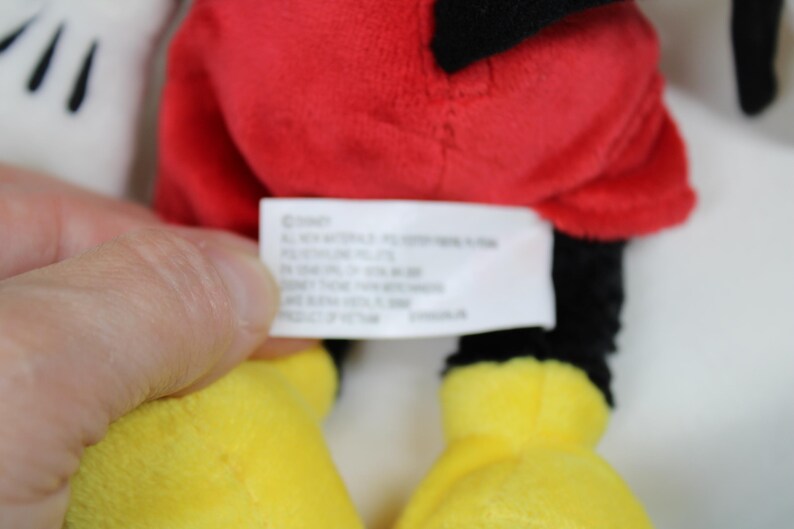 May include: Close-up of a Mickey Mouse plush toy. The toy features a red shorts, black legs, yellow feet, and white gloves. A tag with text is visible.