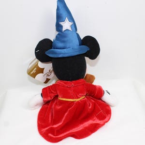 May include: A plush Mickey Mouse figure dressed as a sorcerer, wearing a blue hat with white stars and a red robe. The figure has black ears, white gloves, and a gold belt. A Disney tag is attached.