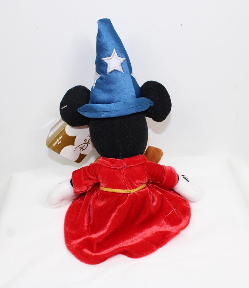May include: A plush Mickey Mouse figure dressed as a sorcerer, wearing a blue hat with white stars and a red robe. The figure has black ears, white gloves, and a gold belt. A Disney tag is attached.