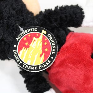 May include: Close-up of a Mickey Mouse plush toy. The toy has black fur, red shorts, white gloves, and a black tail. An "Authentic Original Disney Theme Parks" tag is attached.
