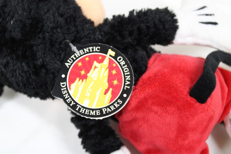 May include: Close-up of a Mickey Mouse plush toy. The toy has black fur, red shorts, white gloves, and a black tail. An "Authentic Original Disney Theme Parks" tag is attached.