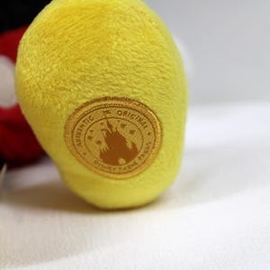 May include: Close-up of a yellow plush toy foot with a gold-colored circular patch. The patch reads "AUTHENTIC ORIGINAL DISNEY THEME PARKS" around a silhouette of a castle. The toy's red and black parts are visible in the background.