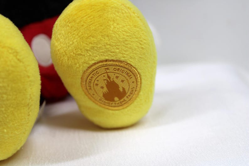 May include: Close-up of a yellow plush toy foot with a gold-colored circular patch. The patch reads "AUTHENTIC ORIGINAL DISNEY THEME PARKS" around a silhouette of a castle. The toy's red and black parts are visible in the background.