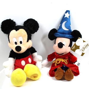 May include: Two plush Mickey Mouse figures. One is in his classic outfit with red shorts and yellow shoes. The other is dressed as a sorcerer, wearing a blue hat with a crescent moon and stars, and a red robe. Two yellow plush balls are in front.
