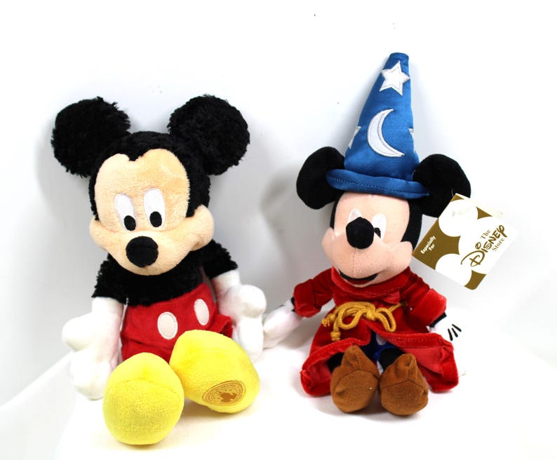 May include: Two plush Mickey Mouse figures. One is in his classic outfit with red shorts and yellow shoes. The other is dressed as a sorcerer, wearing a blue hat with a crescent moon and stars, and a red robe. Two yellow plush balls are in front.