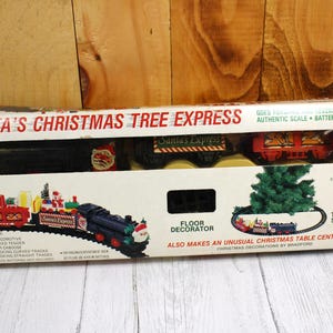 May include: A boxed Christmas train set labeled "Santa's Christmas Tree Express." The box features illustrations of a train, a Christmas tree, and the text "Floor Decorator." The set includes a locomotive, gift-packed tender, and tracks.