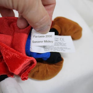 May include: A plush Sorcerer Mickey Mouse doll from Fantasia 2000. The doll features a red velvet robe, a blue hat, and brown shoes. The tag reads "Fantasia 2000 Sorcerer Mickey".