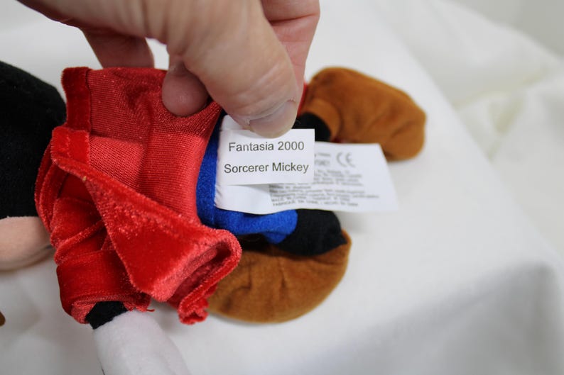 May include: A plush Sorcerer Mickey Mouse doll from Fantasia 2000. The doll features a red velvet robe, a blue hat, and brown shoes. The tag reads "Fantasia 2000 Sorcerer Mickey".