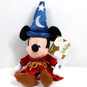 May include: A plush Mickey Mouse dressed as a sorcerer, wearing a blue hat with a white crescent moon and star. He has a red robe with a gold rope belt, white gloves, and brown shoes. A Disney Store tag is attached.