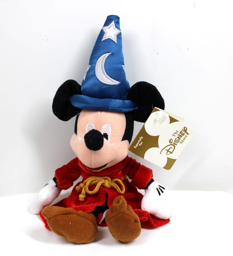 May include: A plush Mickey Mouse dressed as a sorcerer, wearing a blue hat with a white crescent moon and star. He has a red robe with a gold rope belt, white gloves, and brown shoes. A Disney Store tag is attached.