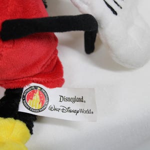 May include: A plush toy featuring a red body, black legs and arms, and a yellow foot. A white glove with black details is visible. The tag reads "Disneyland Walt Disney World".