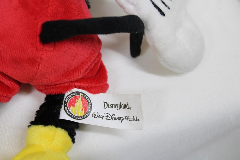 May include: A plush toy featuring a red body, black legs and arms, and a yellow foot. A white glove with black details is visible. The tag reads "Disneyland Walt Disney World".