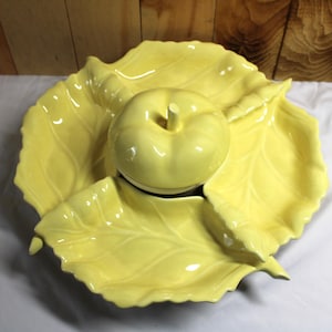 May include: A bright yellow ceramic serving dish shaped like a large leaf with a central apple-shaped bowl. The glossy finish highlights the embossed veins of the leaf sections and the smooth, rounded contours of the central apple container.
