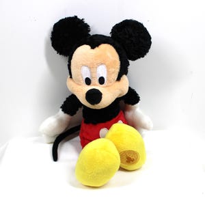 May include: A plush Mickey Mouse toy, featuring black ears, a black body, red shorts, and yellow feet. The toy has white gloves and a smiling face. The toy is a classic Disney character.