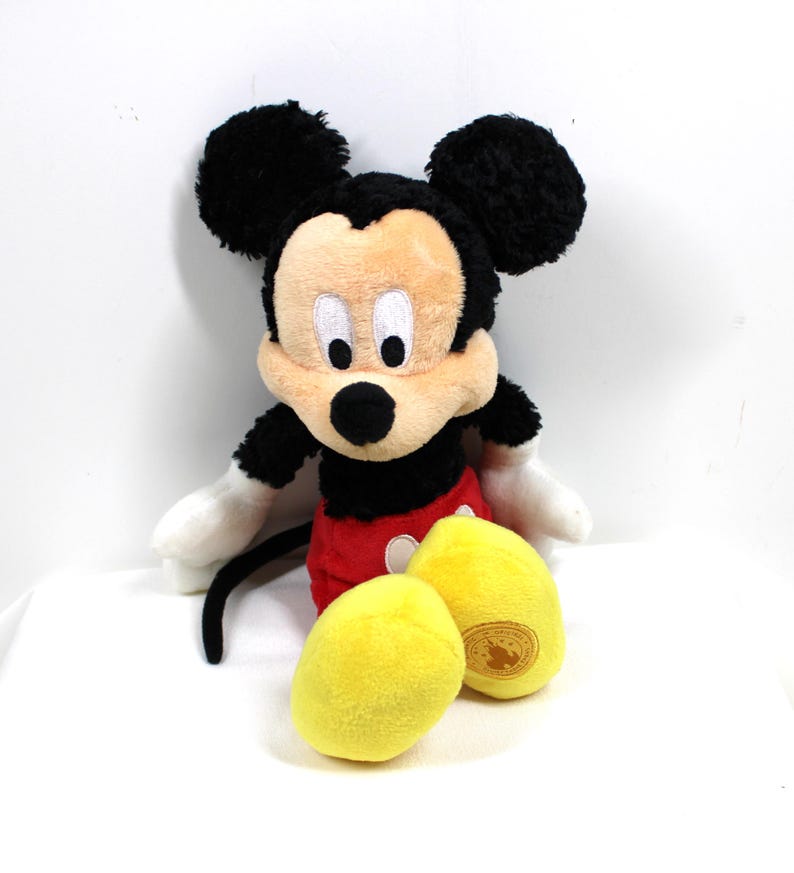 May include: A plush Mickey Mouse toy, featuring black ears, a black body, red shorts, and yellow feet. The toy has white gloves and a smiling face. The toy is a classic Disney character.