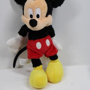 May include: A plush Mickey Mouse toy. The iconic character has a black head with large, round ears, a tan face, and a black nose. He wears red shorts with white buttons, white gloves, and yellow shoes. The toy is soft and cuddly.