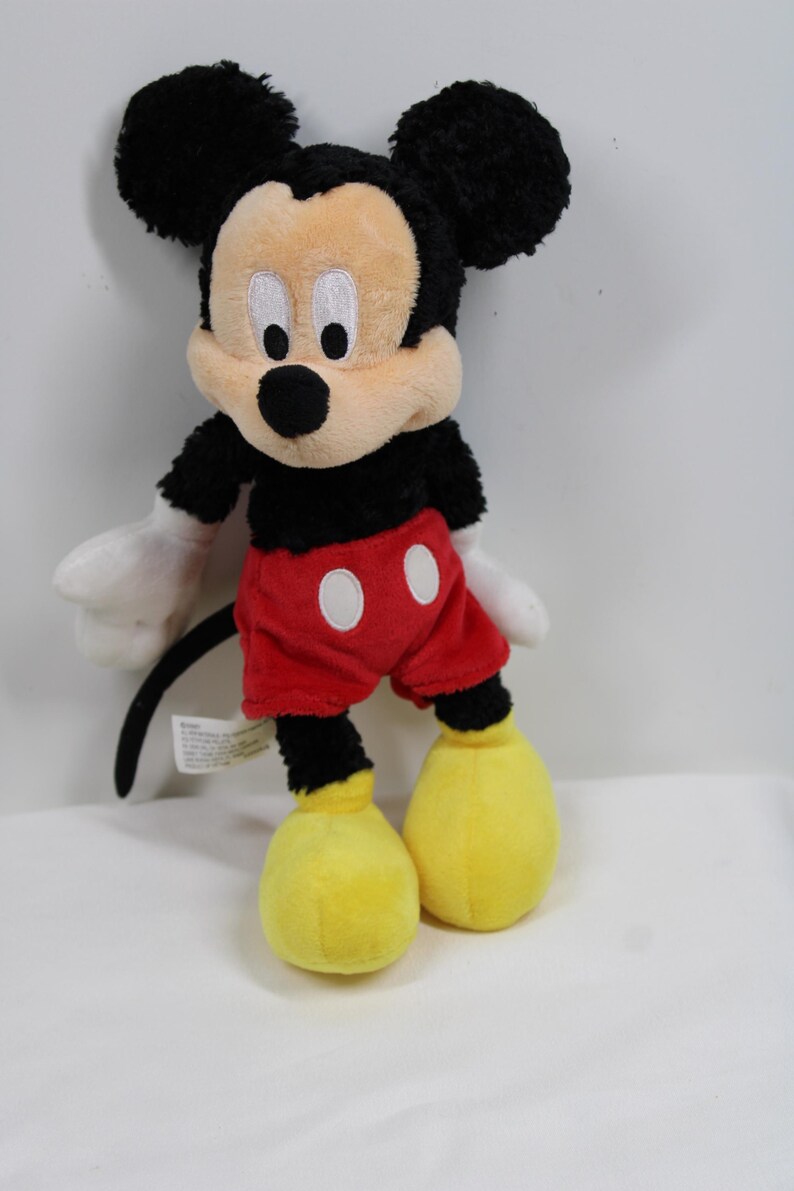 May include: A plush Mickey Mouse toy. The iconic character has a black head with large, round ears, a tan face, and a black nose. He wears red shorts with white buttons, white gloves, and yellow shoes. The toy is soft and cuddly.