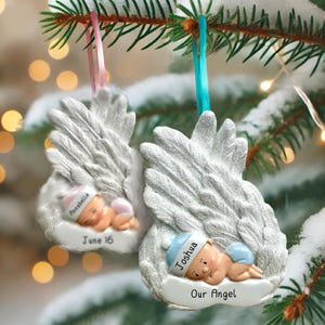 May include: Two angel wing ornaments with sleeping baby figures. One ornament has the name "Annabella" and the date "June 16". The other ornament has the name "Joshua" and the text "Our Angel". Both ornaments have pink and blue ribbons for hanging.