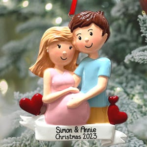 Puede incluir: Un adorno navideño personalizado con una pareja de dibujos animados, un hombre con cabello castaño y una camisa azul, y una mujer con cabello rubio que lleva un vestido rosa. La mujer está embarazada y la pareja se abraza. El adorno tiene una pancarta con el texto "Simon & Annie Navidad 2023".