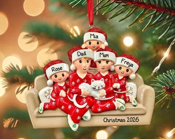 Könnte beinhalten: Ein Weihnachtsbaumschmuck mit einer vierköpfigen Familie und einer Katze auf einem beigen Sofa. Die Figuren tragen rote Schlafanzüge mit weißen Schneeflockenmustern und rote Weihnachtsmützen. Die Mützen sind mit Mia, Dad, Mum und Freya beschriftet. Der Sockel trägt die Aufschrift Christmas 2026.